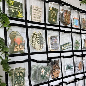 May include: A white hanging organizer with clear plastic pockets containing various rectangular and die-cut stickers. The stickers feature nature-themed illustrations and text including "less screen more green," "inhale," "love your journey," and "nature is medicine."