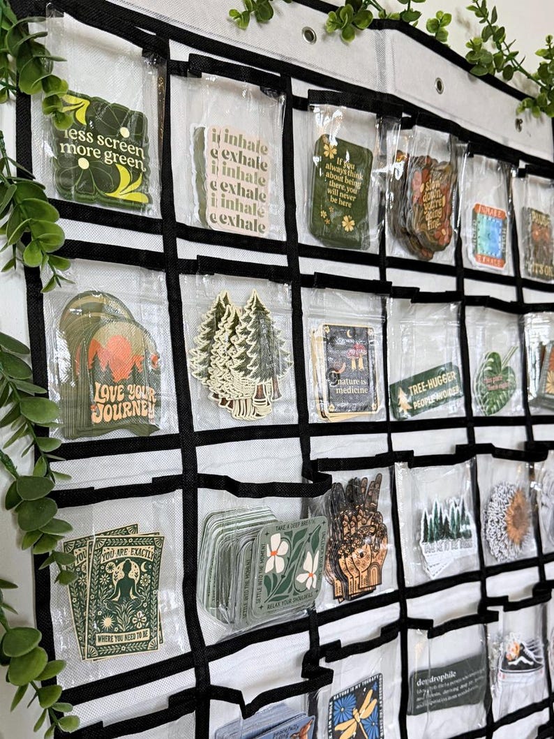 May include: A white hanging organizer with clear plastic pockets containing various rectangular and die-cut stickers. The stickers feature nature-themed illustrations and text including "less screen more green," "inhale," "love your journey," and "nature is medicine."