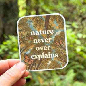 May include: A square sticker with rounded corners featuring a low-angle view of tree branches against a blue sky. The text "nature never explains" is printed in white in the center, surrounded by three orange and black monarch butterflies.
