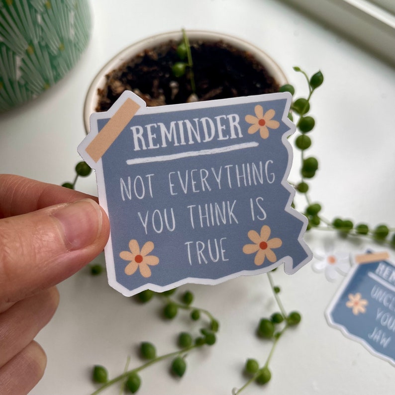 May include: A blue sticker with a white border and rounded corners. The sticker has a white and orange flower on each side and says "Reminder: Not everything you think is true".