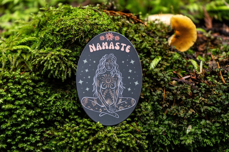 May include: A black oval sticker with a gold outline and a white illustration of a woman sitting in a yoga pose. The woman has long hair and is surrounded by stars and other celestial symbols. The sticker says "NAMASTE" in pink letters.