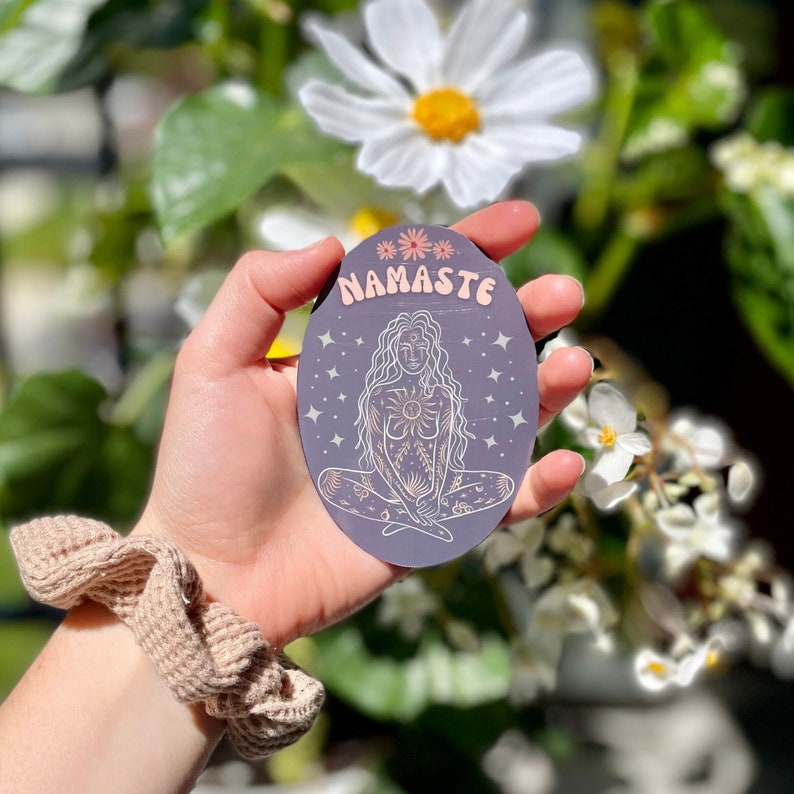 May include: A purple and white sticker with a woman sitting in a yoga pose. The sticker has the word "Namaste" written on it.