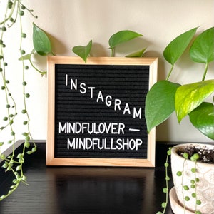 May include: A black letter board with a wooden frame sits on a black surface. The board displays the text "INSTAGRAM - MINDFULOVER - MINDFULLSHOP".  A green potted plant with yellow leaves is in the background.