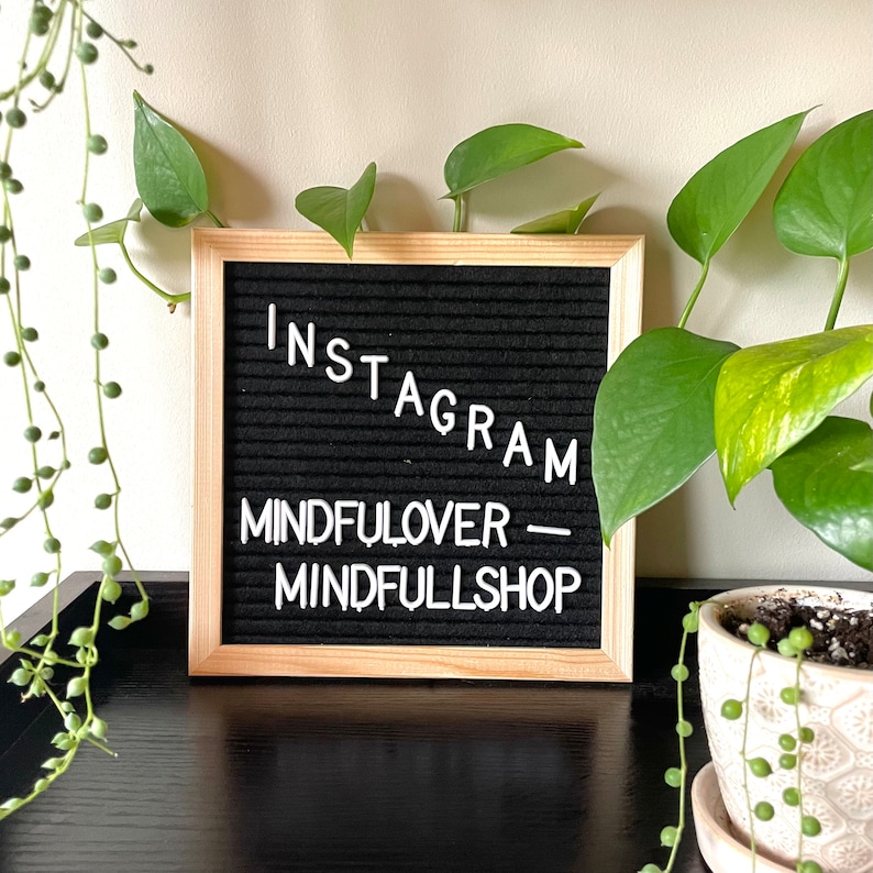 May include: A black letter board with a wooden frame sits on a black surface. The board displays the text "INSTAGRAM - MINDFULOVER - MINDFULLSHOP".  A green potted plant with yellow leaves is in the background.
