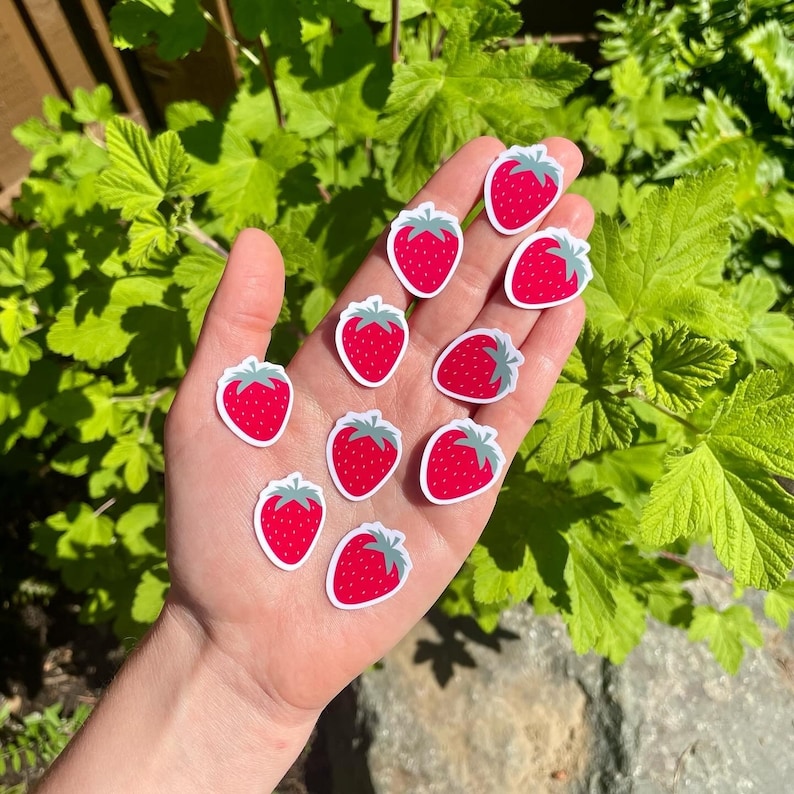 May include: A hand holding a variety of red strawberry stickers. The stickers are all different sizes and shapes, and some have green leaves.