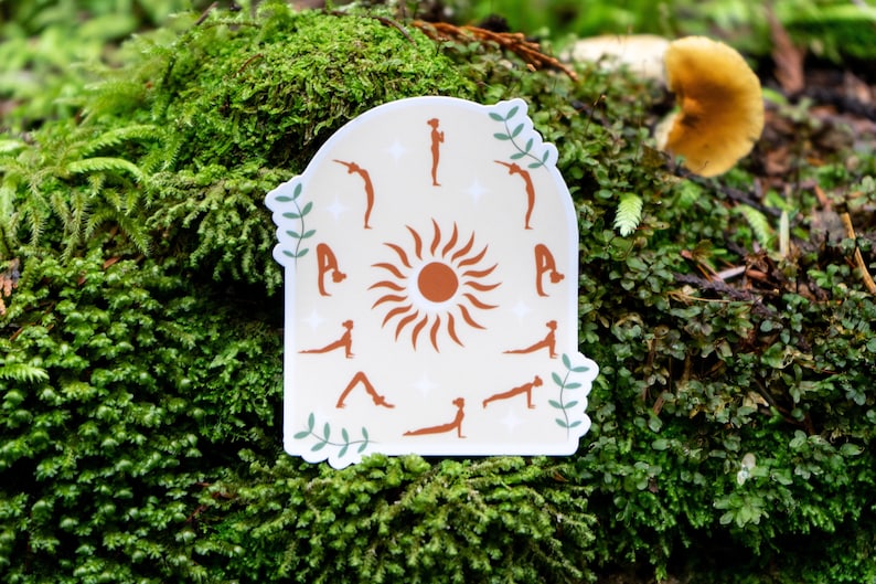 May include: A sticker with a white background and a brown sun in the center. The sun has rays that extend outward. Around the sun are 12 brown silhouettes of people in different yoga poses. The sticker has a rounded arch shape at the top and a green leafy design at the bottom.
