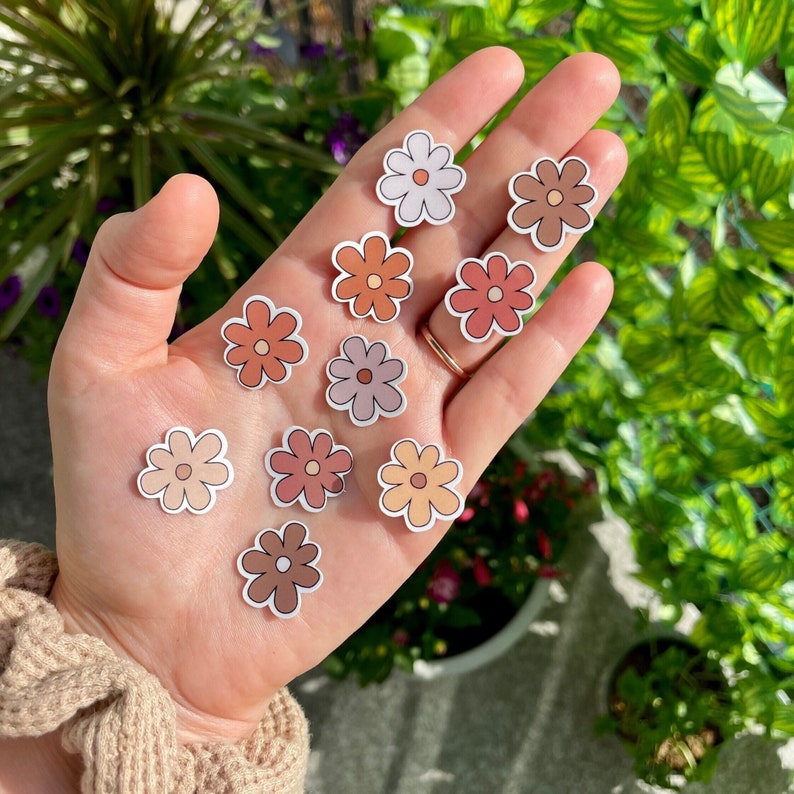 Peut inclure: Une main tenant une vari&eacute;t&eacute; d'autocollants de fleurs color&eacute;s. Les autocollants sont dans des tons de rose, orange et marron. Ils sont tous en forme de marguerite simple.