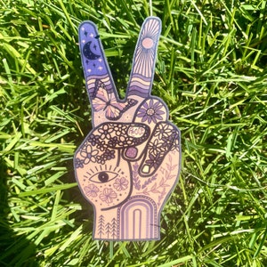 May include: A die-cut sticker shaped like a hand making a peace sign, featuring a light peach background with purple and dark blue line art. The design includes a crescent moon, sun rays, a butterfly, flowers, an eye, and the word Peace.