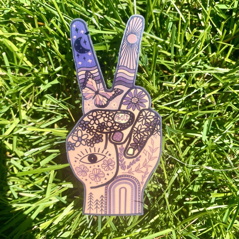 May include: A die-cut sticker shaped like a hand making a peace sign, featuring a light peach background with purple and dark blue line art. The design includes a crescent moon, sun rays, a butterfly, flowers, an eye, and the word Peace.
