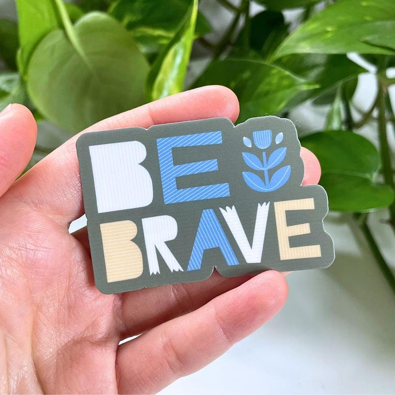 May include: A sticker with the words "BE BRAVE" in white, blue, and tan lettering. The sticker has a floral design and is on a gray background. The sticker is held in a hand.