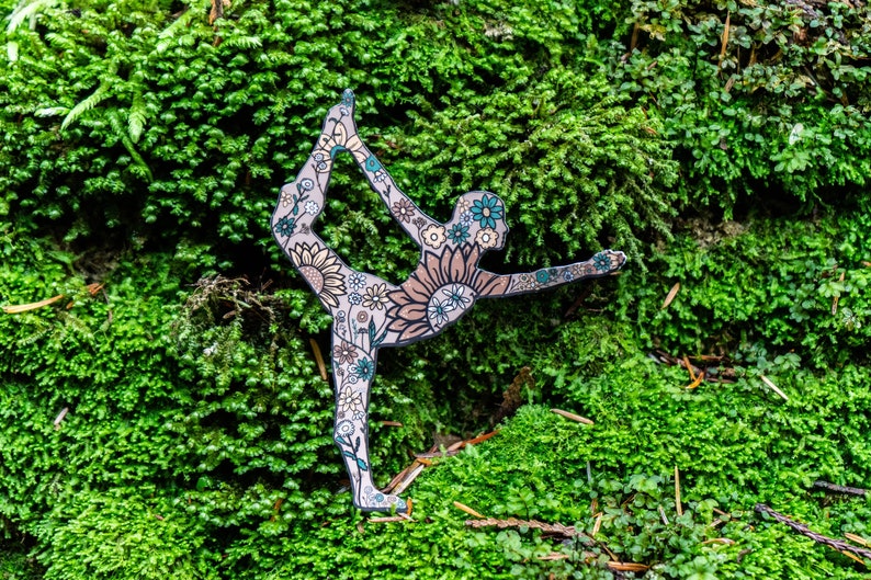 May include: A cutout of a person in a yoga pose, decorated with floral patterns, is placed on a bed of green moss.