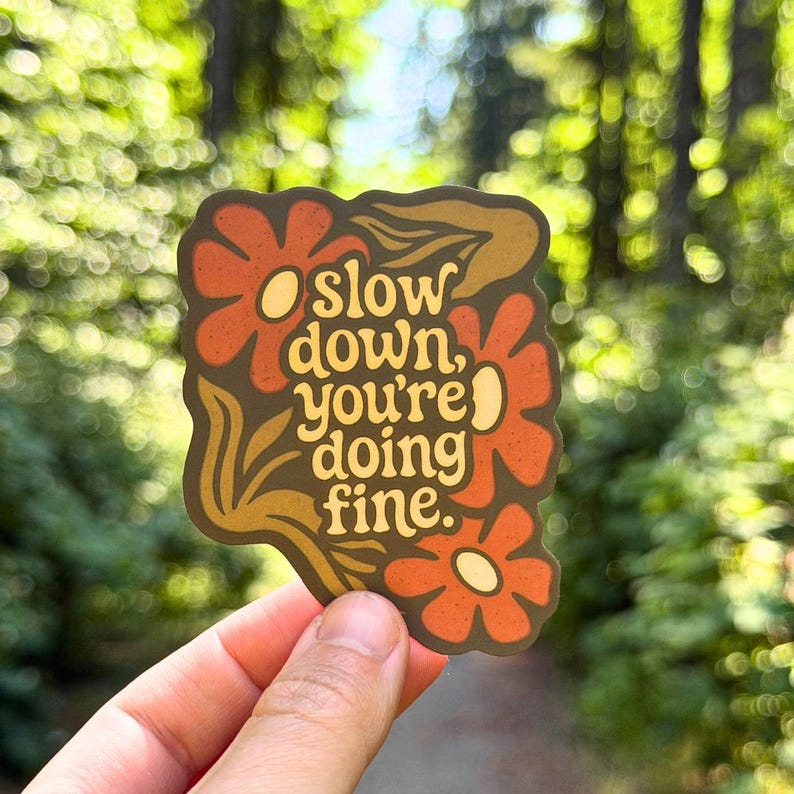 May include: A die-cut sticker with a dark brown background featuring orange flowers and yellow centers. The text in the center reads, "slow down, you're doing fine." The sticker has a wavy, organic border shape and a matte finish with a textured appearance.