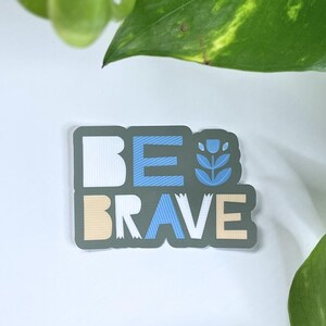 May include: A rectangular sticker with the words "BE BRAVE" in white, blue, and tan. The sticker has a dark gray background and a floral design. The sticker is on a white surface with green leaves in the background.