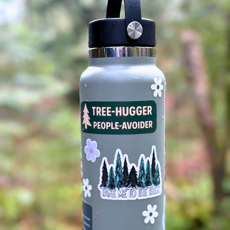 May include: A gray water bottle with a black lid and silver accents. The bottle features stickers with the phrases "TREE-HUGGER PEOPLE-AVOIDER" and "TAKE ME TO THE TREES", along with floral and tree graphics.