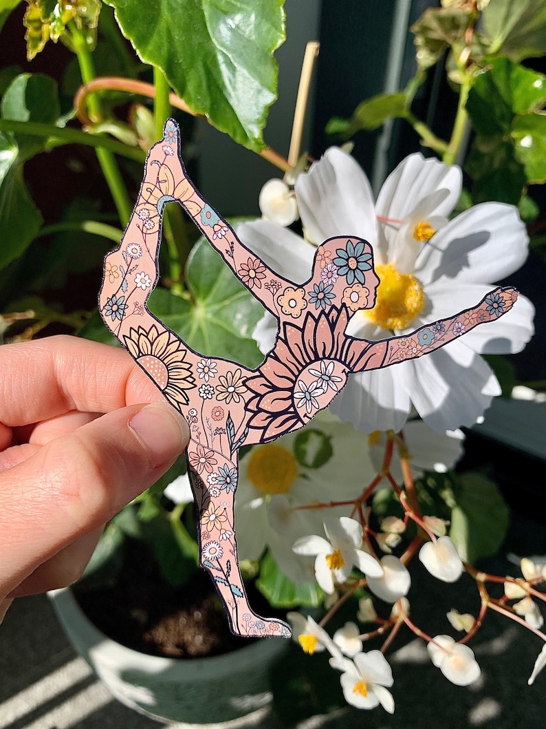 May include: A sticker of a yoga pose with a floral pattern. The pose is a variation of the dancer's pose. The sticker is pink with a floral pattern in shades of yellow, blue, and green.