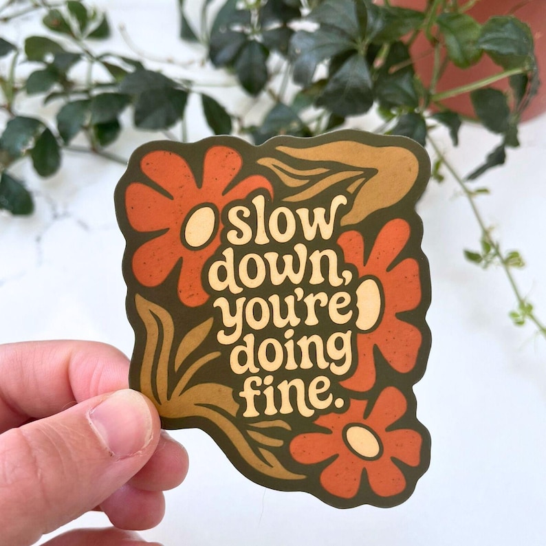 May include: A sticker with a retro floral design in shades of orange, brown, and olive green. The sticker features the text "slow down, you're doing fine" in a cream-coloured font, perfect for adding a touch of positivity to any item.