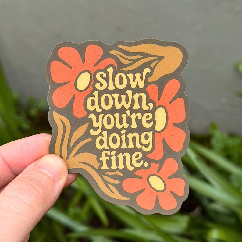 May include: A die-cut sticker with a dark brown background featuring orange flowers with yellow centers and yellow leaves. The text in the center reads, "slow down, you're doing fine." in a rounded, bold, yellow, retro-style font.