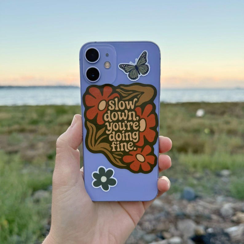 May include: A lilac phone case with a floral design and the text "slow down, you're doing fine." The case also features a butterfly and a small green flower sticker. The phone case is held in a person's hand.