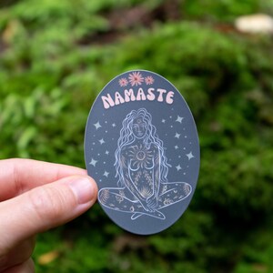 May include: A gray oval sticker with a white outline and a pink and white illustration of a woman sitting in a yoga pose. The sticker has the word "Namaste" written in pink letters at the top.