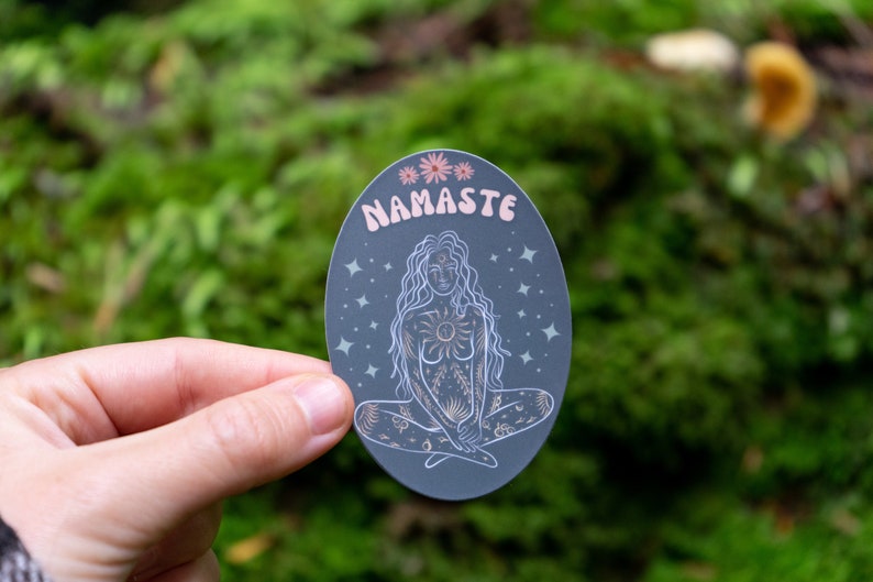 May include: A gray oval sticker with a white outline and a pink and white illustration of a woman sitting in a yoga pose. The sticker has the word "Namaste" written in pink letters at the top.