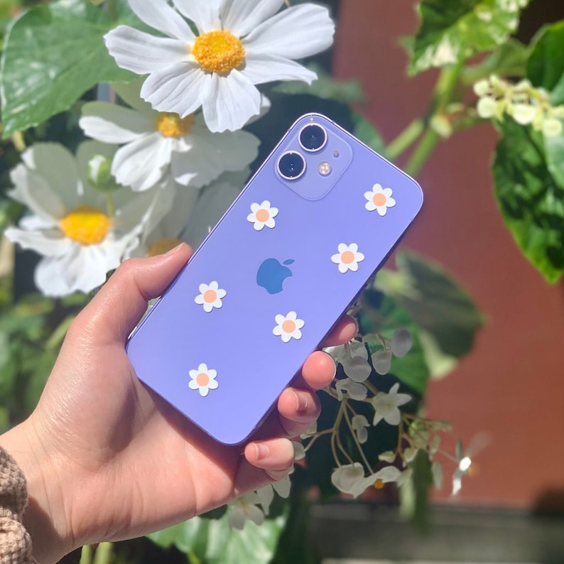 May include: A purple iPhone case with white daisy stickers. The case has a camera bump with three lenses.