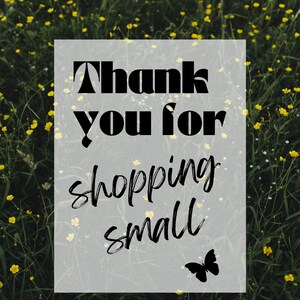 May include: A white sign with black text that reads "Thank you for shopping small" with a black butterfly silhouette in the bottom right corner. The sign is in front of a field of green grass and yellow flowers.