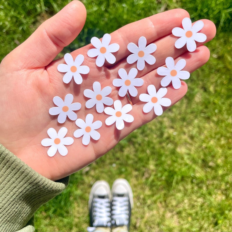 May include: A hand holding a collection of white daisy stickers with orange centres. The stickers are arranged in a semi-circle shape.