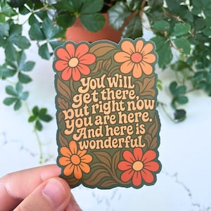 Może przedstawiać: Prostokątna naklejka z kwiatowym wzorem i napisem "You will get there, but right now you are here. And here is wonderful." Naklejka przedstawia pomarańczowe i czerwone kwiaty na brązowym i zielonym tle.