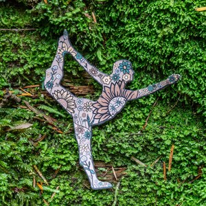 May include: A paper cutout of a person in a yoga pose, decorated with floral patterns in shades of green, blue, and brown. The cutout is placed on a bed of green moss.