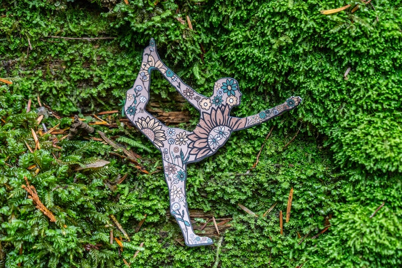 May include: A paper cutout of a person in a yoga pose, decorated with floral patterns in shades of green, blue, and brown. The cutout is placed on a bed of green moss.