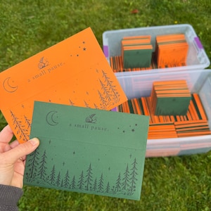May include: Two rectangular envelopes in orange and dark green, each featuring a black graphic of a snail, a crescent moon, stars, and a forest of pine trees. The text "a small pause." is printed on the front of each envelope.