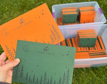 May include: Two rectangular envelopes in orange and dark green, each featuring a black graphic of a snail, a crescent moon, stars, and a forest of pine trees. The text "a small pause." is printed on the front of each envelope.