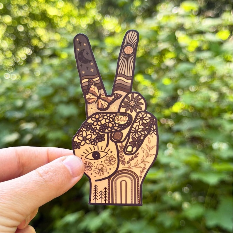 May include: A die-cut sticker shaped like a hand making a peace sign, featuring a warm-toned palette of brown, tan, and cream. The design includes intricate illustrations of a crescent moon, sun, butterfly, flowers, an eye, and the word Peace.