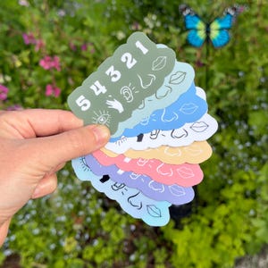 May include: A stack of seven die-cut stickers in various colors including olive green, light blue, white, yellow, pink, purple, and light blue. Each sticker features the numbers 5 4 3 2 1 and white line art icons of an eye, hand, ear, nose, and lips.