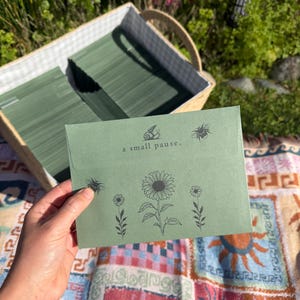 May include: A sage green envelope featuring black line art illustrations of a sunflower, two smaller flowers, two bees, and a snail. The text "a small pause." is printed in black on the flap. The envelope is held against a background of a basket.