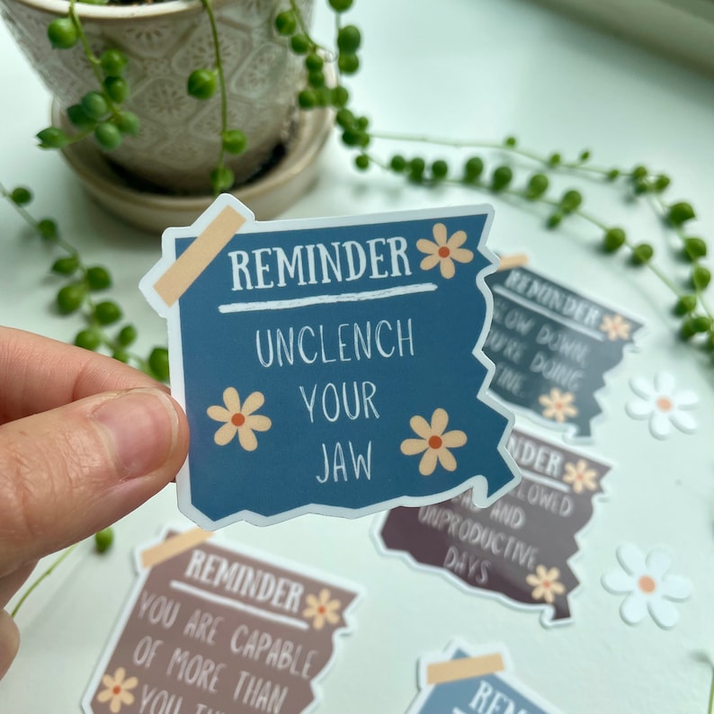 May include: A set of four blue, brown and grey stickers with a white daisy design. The stickers have the text "Reminder" and a short message. The text on the blue sticker reads "Unclench Your Jaw".