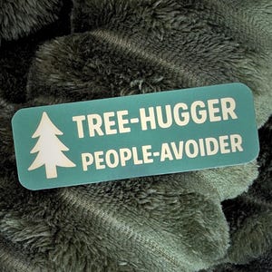 May include: A teal rectangular sticker with rounded corners, featuring a white pine tree graphic and the words "TREE-HUGGER PEOPLE-AVOIDER" in white capital letters. The sticker is set against a backdrop of a soft, textured, olive-green fabric.