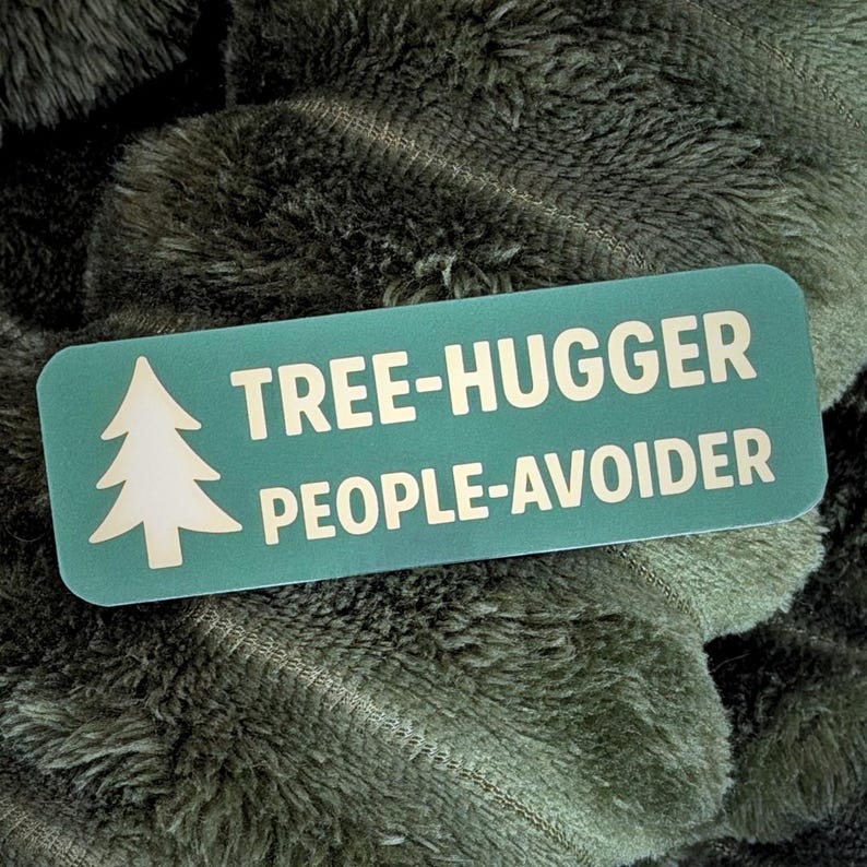 May include: A teal rectangular sticker with rounded corners, featuring a white pine tree graphic and the words "TREE-HUGGER PEOPLE-AVOIDER" in white capital letters. The sticker is set against a backdrop of a soft, textured, olive-green fabric.