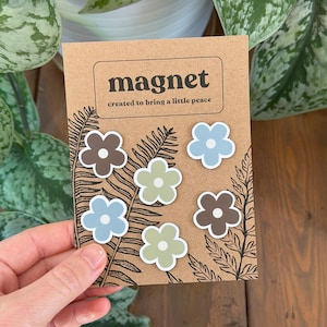 May include: A card of flower-shaped magnets in brown, blue, and green. The card reads "magnet" and "created to bring a little peace." The magnets are a decorative item. The card is held by a person.