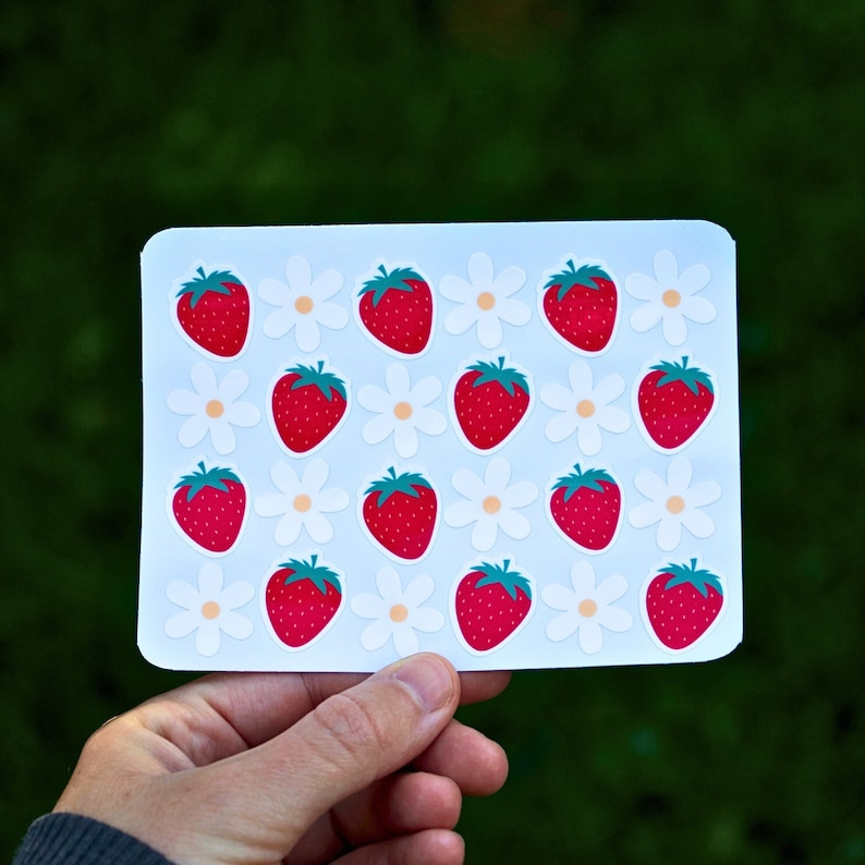 May include: A sheet of white sticker paper with red strawberries and white daisies printed on it. The stickers are arranged in a grid pattern.