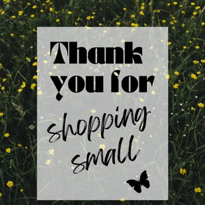 May include: A white rectangle with black text that reads "Thank you for shopping small" with a black butterfly silhouette in the bottom right corner. The rectangle is in front of a field of green grass with yellow flowers.