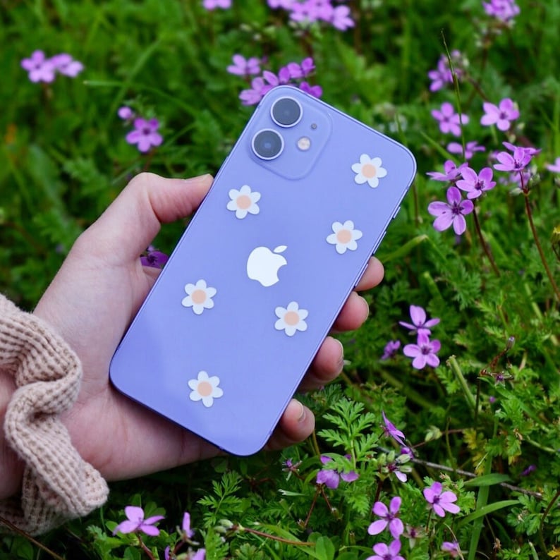 May include: A purple iPhone with a white apple logo and white daisy stickers on the back. The phone is being held in a hand against a background of purple flowers.