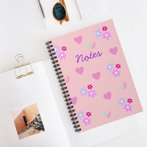 May include: A spiral-bound notebook with a pink cover featuring the word "Notes" in purple script, along with heart and flower illustrations in pink, purple, and blue. The notebook is open, revealing blank pages.