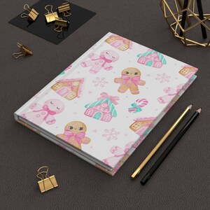 May include: A hardcover journal with a white cover featuring a repeating pattern of gingerbread men, gingerbread houses, and snowflakes in pink, brown, and teal. Gold and black pencils are next to the journal.