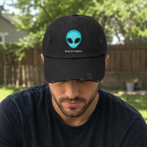 Op de afbeelding: Zwarte distressed baseball cap met een lichtblauw alien gezicht met zwarte ogen. De tekst "Area 51 Reject" staat onder het alien ontwerp. De cap heeft een voorgebogen klep en een versleten, vintage look.