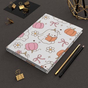 May include: A closed journal with a white cover featuring a repeating pattern of cartoon pumpkins, flowers, bows, and stars. A cartoon ghost is reading a book. Gold and black pencils and gold binder clips are on the dark gray surface.