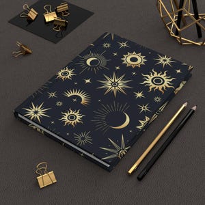 May include: A dark blue journal with a celestial design featuring gold suns, stars, and crescent moons. The notebook is open, revealing white pages. Gold binder clips and pencils are arranged around the journal, suggesting a desk or workspace.