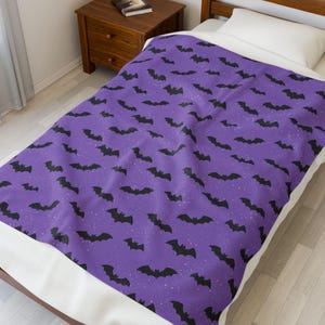 Puede incluir: Una manta morada con un patrón de murciélagos negros y pequeños puntos blancos. La manta está sobre una cama, con una almohada blanca en la parte superior. El diseño tiene un tema de Halloween o de miedo.