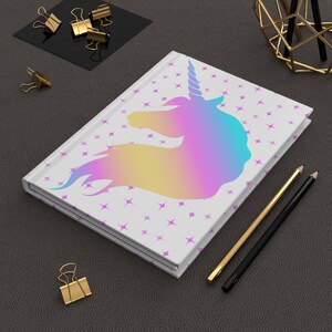 May include: A white journal with a unicorn design in shades of blue, purple, and yellow. The journal has a star pattern and is accompanied by gold and black pencils and gold binder clips.