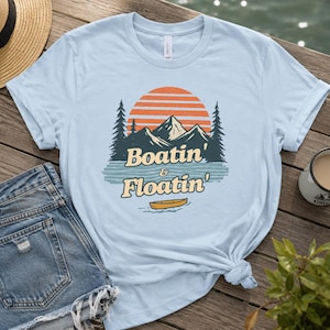Puede incluir: Una camiseta azul claro con un gráfico de una cordillera, una puesta de sol y un pequeño bote amarillo sobre el agua. El texto "Boatin' & Floatin'" está impreso en una fuente retro debajo de las montañas y los pinos de color verde oscuro.